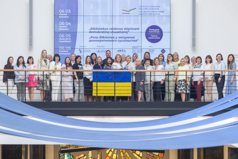 Didelė grupė žmonių išskleidę Ukrainos vėliavą stovi ant tiltelio forumo programos fone