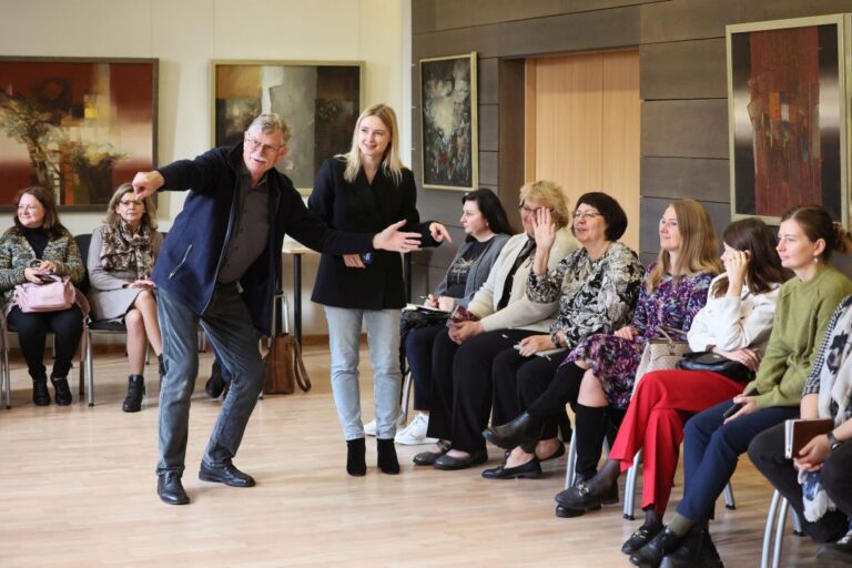 Vyras pritūpęs kažką rodo ranka mokymų dalyvių auditorijoje bibliotekos salės aplinkoje