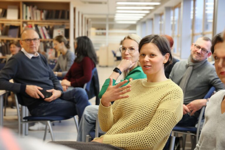 Grupė žmonių sėdi neformaliai ir susidomėję bibliotekos skaityklos aplinkoje