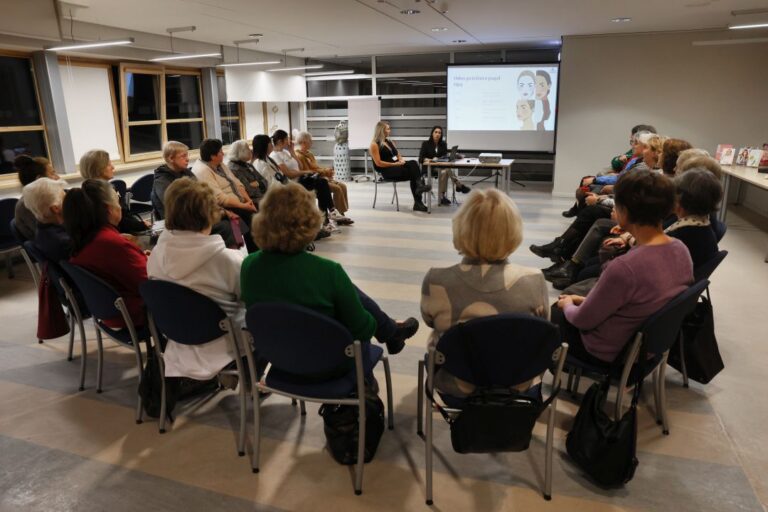 Grupė žmonių sėdi ratu bibliotekos skaityklos fone