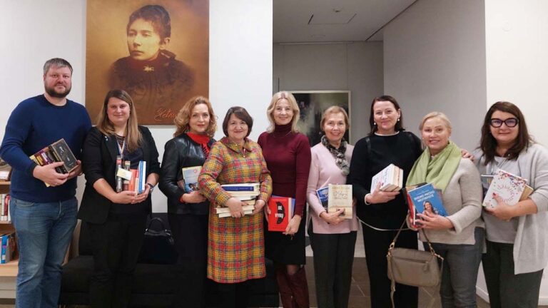 Grupė vyrų ir moterų su knygomis rankose stovi bibliotekos aplinkoje