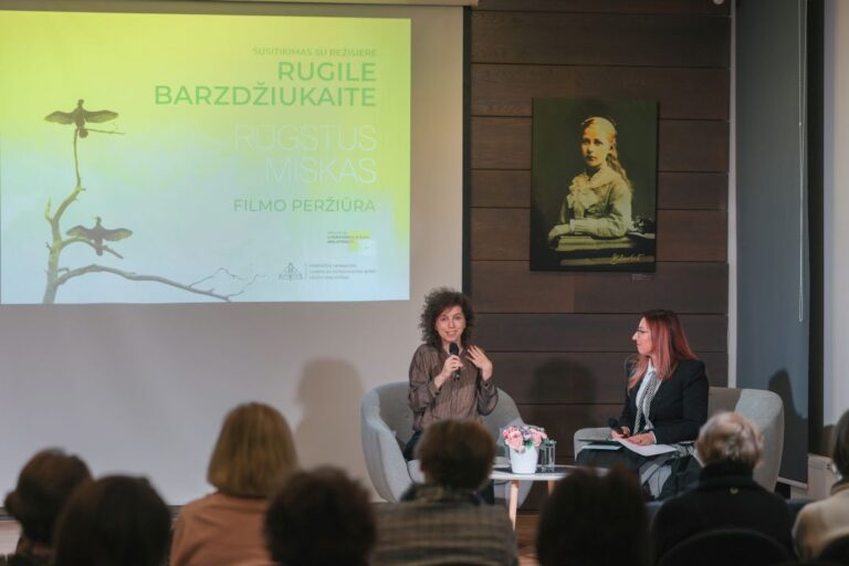 Dvi moterys kalbasi priešais klausytojų auditoriją bibliotekos salėje