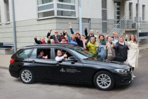 Grupė žmonių iškėlę nykščius šalia juodo automobilio bibliotekos pastato fone 