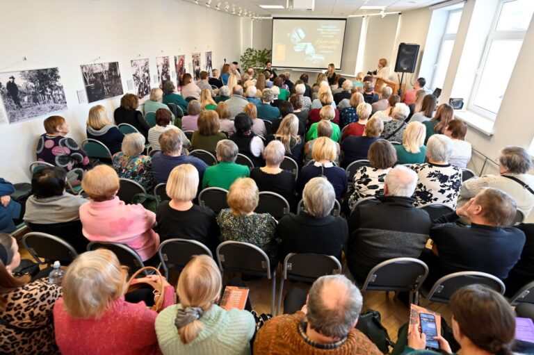 Bibliotekos salė, pilna sėdinčių žmonių ir priekyje sėdintis kunigas Benas Lyris su kalbintoja